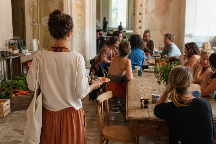 A newcomer holding a breakfast plate beside a busy communal table with an empty chair